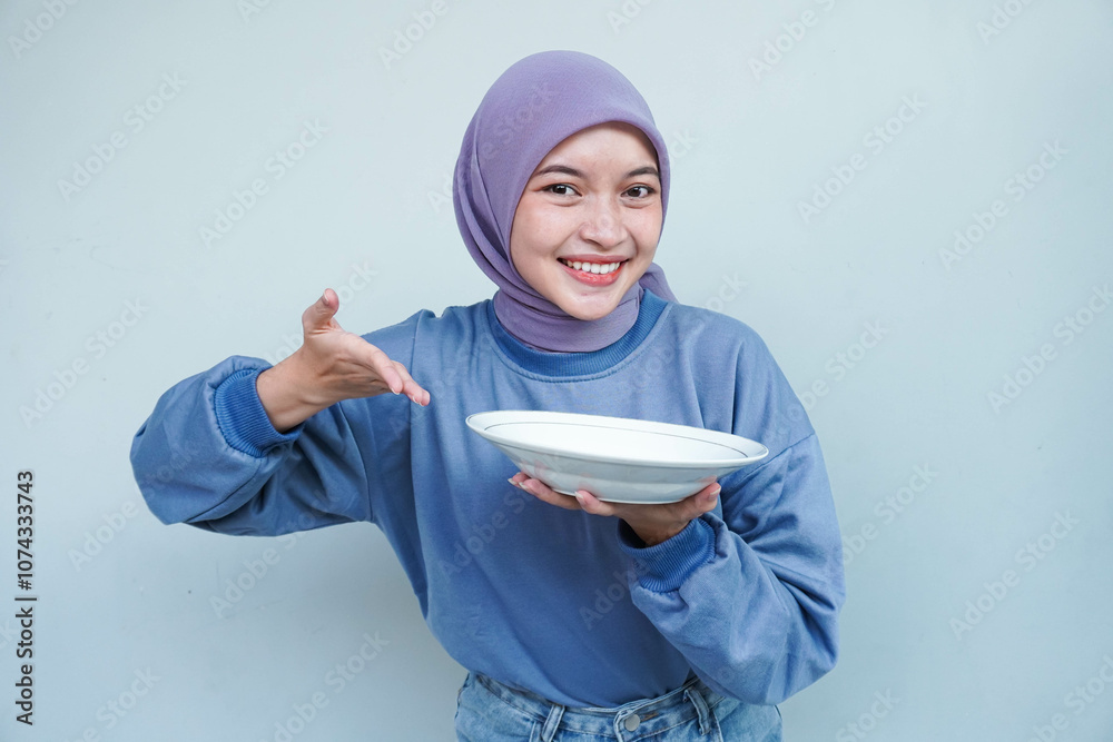 A smiling Asian Muslim woman is fasting and hungry and holding and pointing to a plate. A smiling Asian Muslim woman is fasting and hungry and holding and pointing to a plate.