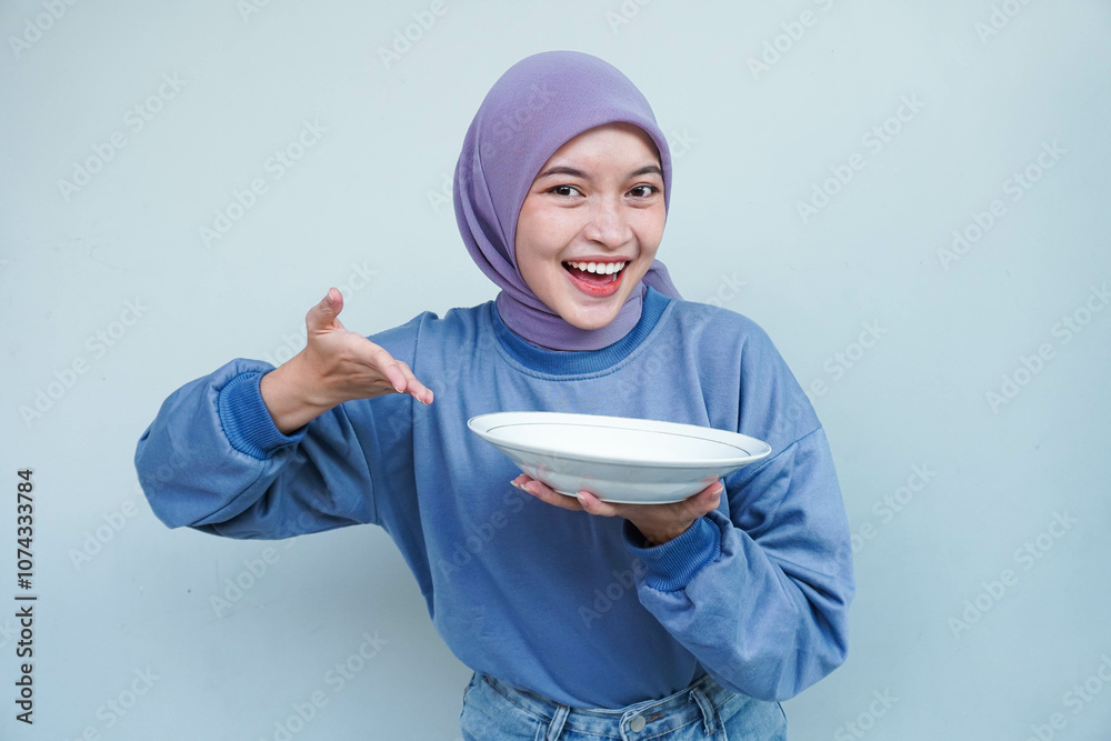 A smiling Asian Muslim woman is fasting and hungry and holding and pointing to a plate. A smiling Asian Muslim woman is fasting and hungry and holding and pointing to a plate.