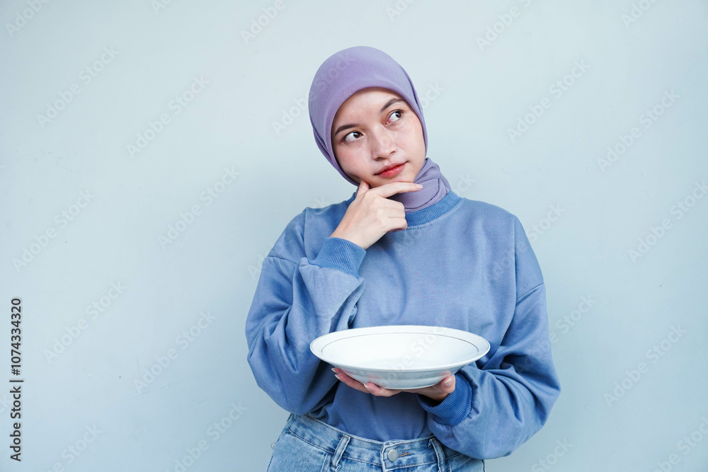 Asian Muslim woman is fasting and hungry and holding cutlery while looking aside thinking about what to eat with empty plate. Asian Muslim woman is fasting and hungry and holding cutlery while looking aside thinking about what to eat with empty plate.