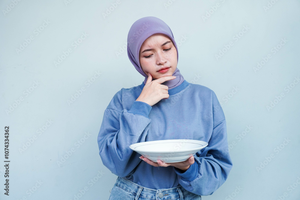 Asian Muslim woman is fasting and hungry and holding cutlery while looking a plate thinking about what to eat with empty plate. Asian Muslim woman is fasting and hungry and holding cutlery while looking a plate thinking about what to eat with empty plate.