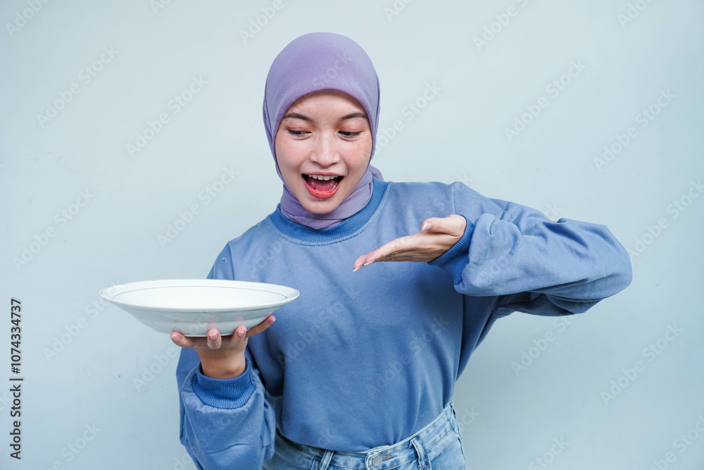 A smiling Asian Muslim woman is fasting and hungry and holding and pointing to a plate. A smiling Asian Muslim woman is fasting and hungry and holding and pointing to a plate.