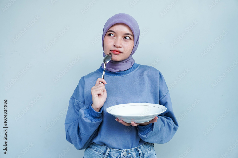 Asian Muslim woman is fasting and hungry and holding cutlery while looking aside thinking about what to eat with empty plate. Asian Muslim woman is fasting and hungry and holding cutlery while looking aside thinking about what to eat with empty plate.