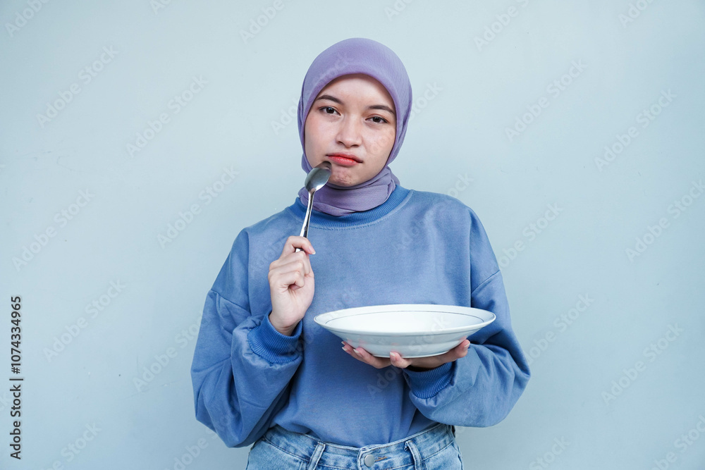 Asian Muslim woman is fasting and hungry and holding cutlery while looking a camera thinking about what to eat with empty plate. Asian Muslim woman is fasting and hungry and holding cutlery while looking a camera thinking about what to eat with empty plate.