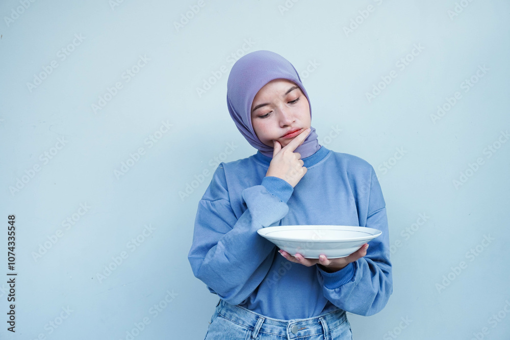 Asian Muslim woman is fasting and hungry and holding cutlery while looking a plate thinking about what to eat with empty plate. Asian Muslim woman is fasting and hungry and holding cutlery while looking a plate thinking about what to eat with empty plate.