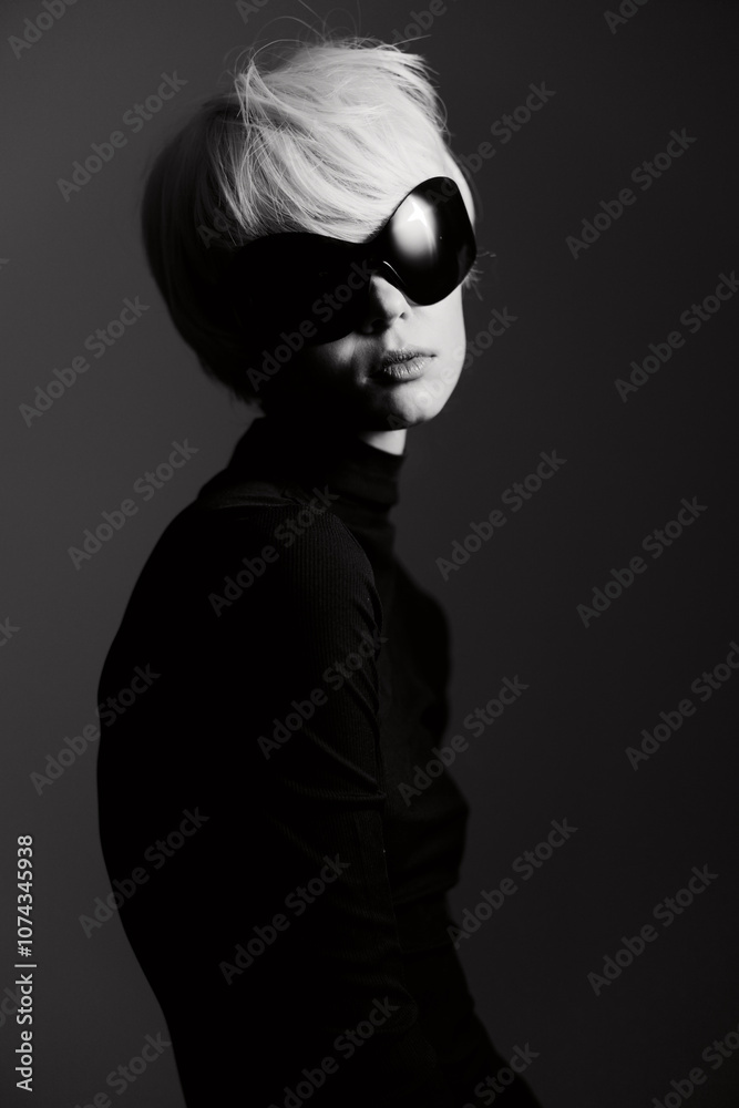 Stylish young woman with short blonde hair and large black sunglasses posing elegantly against a dark background. Fashion and elegance concept. Stylish young woman with short blonde hair and large black sunglasses posing elegantly against a dark background. Fashion and elegance concept.