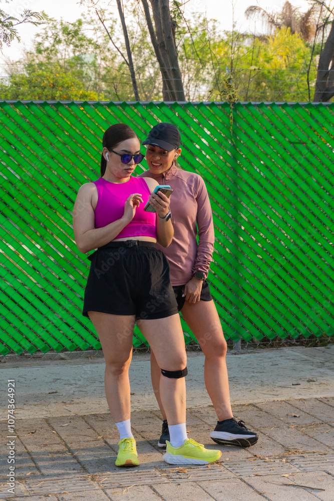Obraz premium Two young Latina women checking smartphone outdoors by green fence