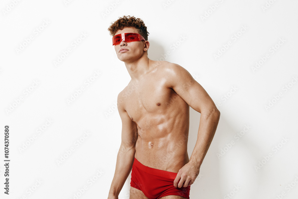 Fototapeta premium Confident young man posing with a muscular build, wearing red swimwear and stylish sunglasses against a plain white background. Summer and fashion concept.