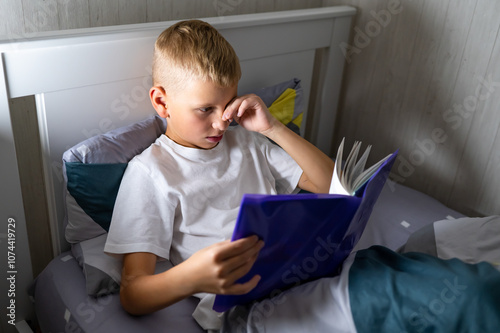 fair-haired teenage boy reads a book before bed and rubs his eyes, wants to sleep photo