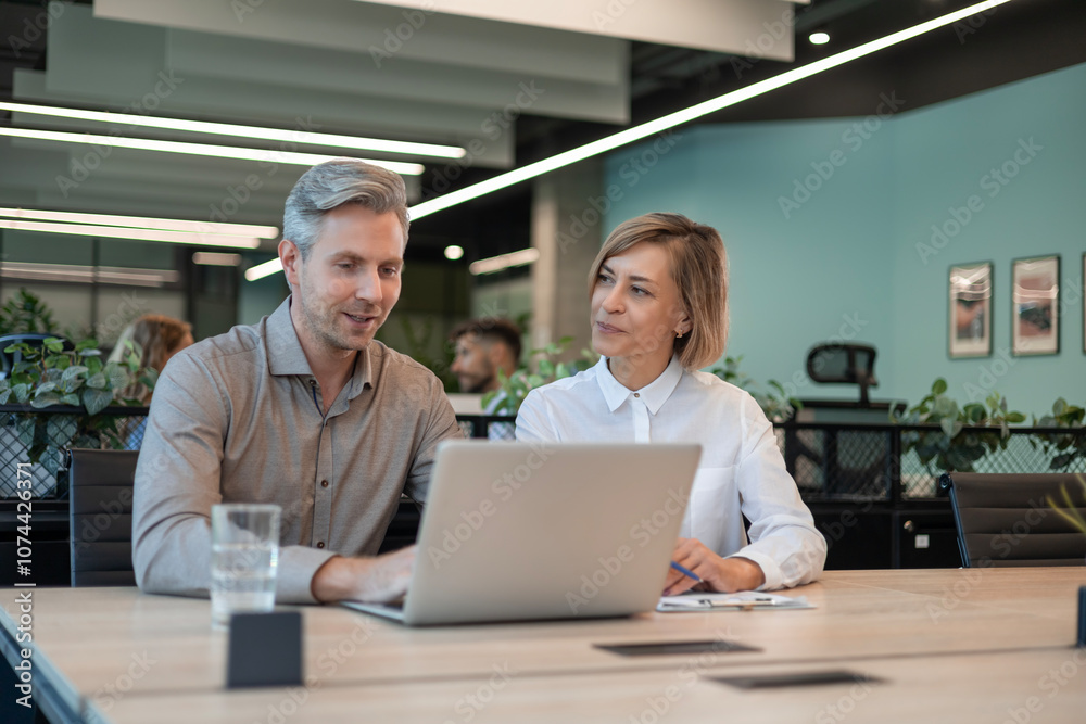 Two business people using laptop on desk discussing strategy company with other employees on background Two business people using laptop on desk discussing strategy company with other employees on background