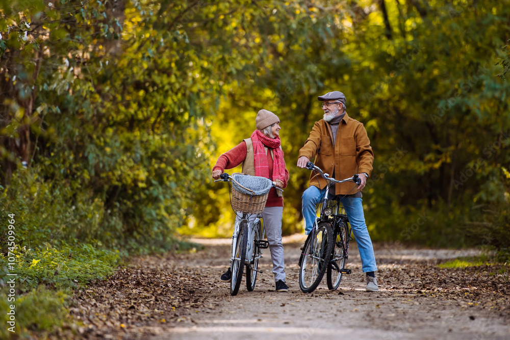 Naklejka premium Elderly couple on a walk in the forest, pushing their bikes side by side. Seniors in love on stroll through autumn nature, enjoying a peaceful moment together.