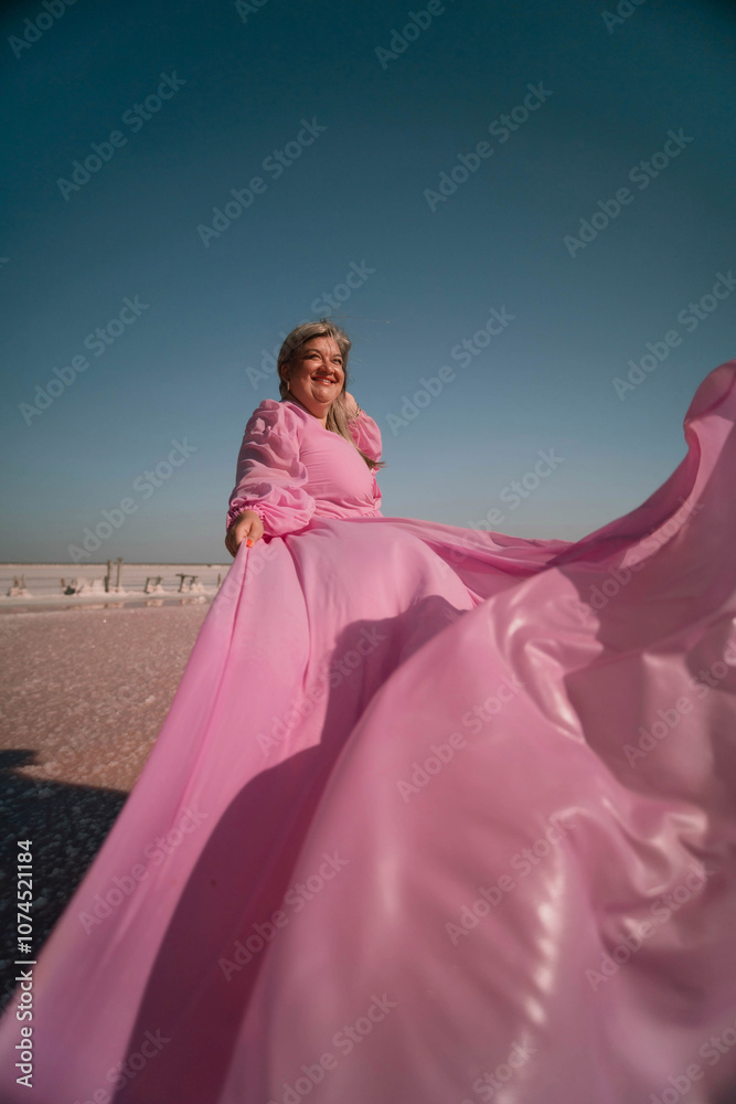 Fototapeta premium Woman Pink Dress Salt Flats - A woman in a pink dress poses on a salt flat with a bright blue sky in the background.