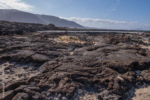 White sand beach with solidified volcanic lava flows, black rocks. Mountains in the background and sky with big white clouds. Caletón Blanco, Lanzarote, Canary Islands, Spain