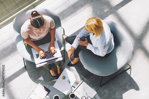 Two diverse business women sit in a large, bright office space and talk together and having a casual meeting. One of the women is taking notes on her laptop. On the table is paper with graphs. 