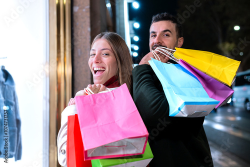 Smiling young couple holding colorful shopping bags, enjoying a night out in the city. They radiate excitement and joy, perfect for concepts related to shopping, lifestyle, and urban experiences photo