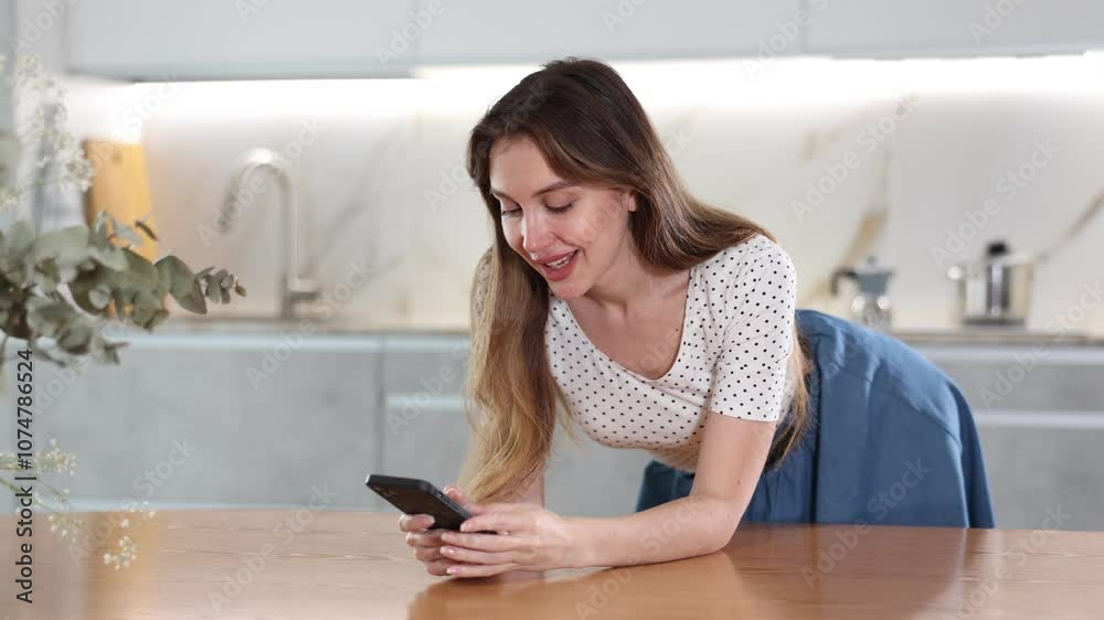 Joyous middle-aged woman leaning on table holding her mobile phone in one hand in the kitchen Joyous middle-aged woman leaning on table holding her mobile phone in one hand in the kitchen