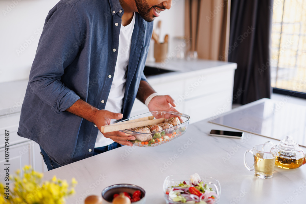 African american man holding baking dish with chicken and vegetables near tea and smartphone in kitchen African american man holding baking dish with chicken and vegetables near tea and smartphone in kitchen