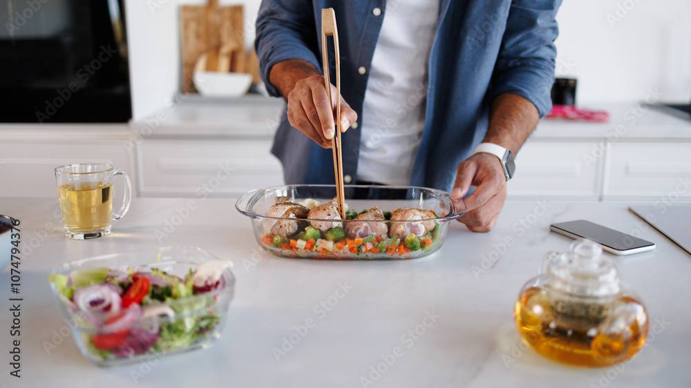 Partial shot of black man holding wooden tongs while cooking chicken and vegetables in baking dish in kitchen Partial shot of black man holding wooden tongs while cooking chicken and vegetables in baking dish in kitchen