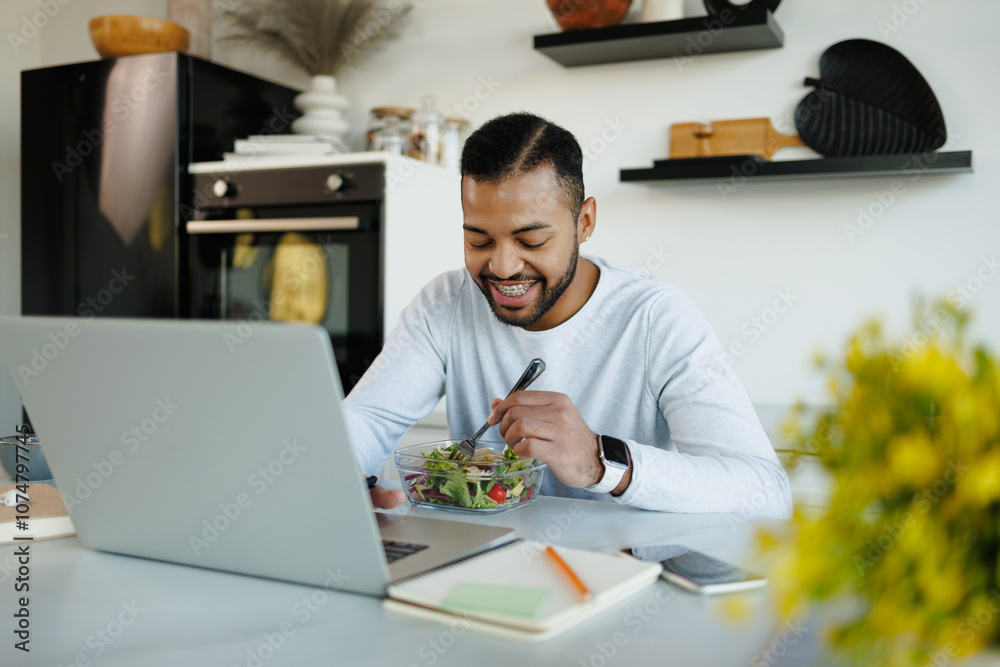 Positive african american freelancer eating fresh salad near devices in kitchen Positive african american freelancer eating fresh salad near devices in kitchen