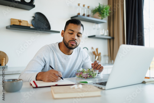 African american freelancer writing on notebook near fresh salad and laptop at home