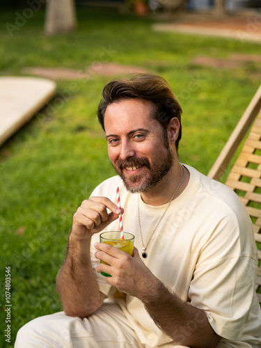 A man enjoys a beverage while sitting on a lounge chair in a green garden. He has a cheerful expression and a striped straw in his glass, surrounded by natural beauty. photo