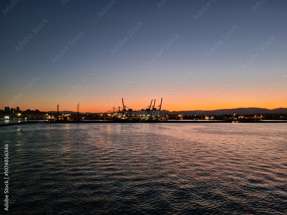 Naklejka premium evocative image of the sun setting behind the sea coast with the solhoutte of the port in the background in Italy