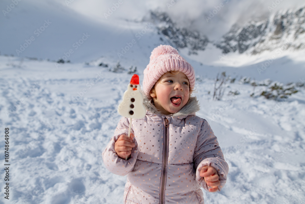 © Halfpoint - Small girl licking a snowman-shaped lollipop, standing in the middle of snowy nature. Genjoying winter holiday in the mountains with family.