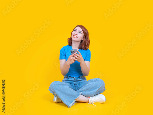 Dreamful woman looking copy space, ful body front view caucasian 20s red bob hair dreamful woman looking copy space wear casual jean blue t-shirt hold use mobile phone isolated on yellow background.