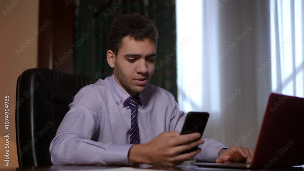 A young man dressed in formal business attire is intently using his smartphone while diligently working at a desk A young man dressed in formal business attire is intently using his smartphone while diligently working at a desk