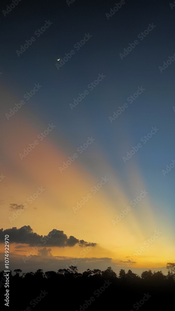 Scenic view of a crescent moon hanging over a colorful twilight sky with warm tones of orange and yellow, casting soft rays of light above a silhouetted tree line. Scenic view of a crescent moon hanging over a colorful twilight sky with warm tones of orange and yellow, casting soft rays of light above a silhouetted tree line.