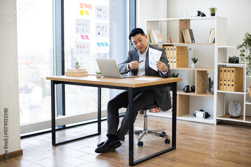 Asian businessman wearing headset in office showing graphs during online video call. Professional setting with laptop, modern interior, and large window. Business attire suggests formal meeting.