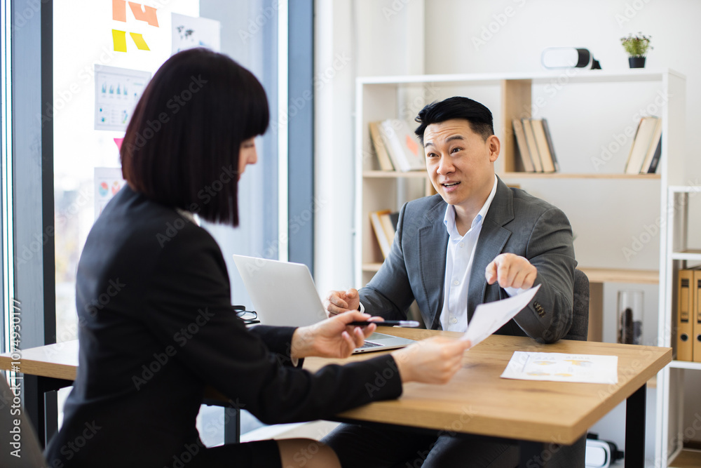 Caucasian businesswoman discussing report with Asian businessman boss in modern office. Business environment includes laptop and documents reflecting teamwork and collaboration. Caucasian businesswoman discussing report with Asian businessman boss in modern office. Business environment includes laptop and documents reflecting teamwork and collaboration.