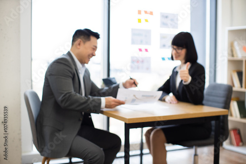 Blurred background. Asian businessman presents report to Caucasian businesswoman in office setting. Both individuals display positive and professional demeanor.