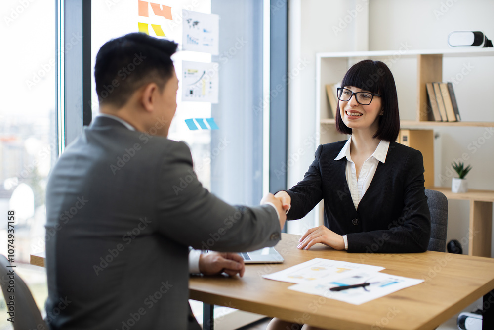 Caucasian woman and Asian man in business attire shaking hands in office. Concept of successful business agreement collaboration. Sunlight streaming through windows enhances atmosphere. Caucasian woman and Asian man in business attire shaking hands in office. Concept of successful business agreement collaboration. Sunlight streaming through windows enhances atmosphere.