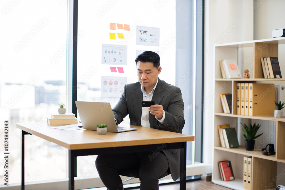 Fototapeta premium Asian businessman in formal suit sitting at office desk, using laptop and bank card for online payment. Bright office setting with bookshelves and window view enhances professional atmosphere.