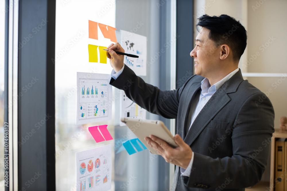 Asian businessman in formal suit analyzing graphs and charts on panoramic window using digital tablet. He is focused on data visualization and office productivity. Asian businessman in formal suit analyzing graphs and charts on panoramic window using digital tablet. He is focused on data visualization and office productivity.