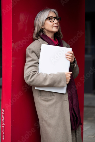 An older woman in a beige coat stands by a bright red wall, holding a white folder while looking thoughtfully into the distance photo