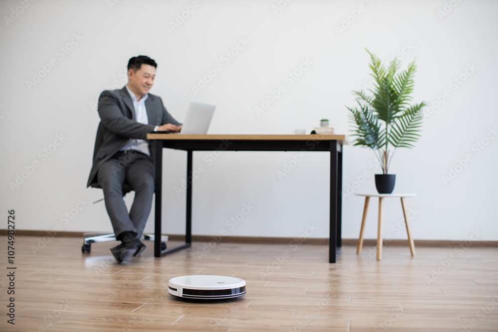 Asian businessman sitting at table using laptop in modern office space, while robot vacuum cleans floor. Scene depicts efficient, tech-savvy workspace with clean, minimalist design.