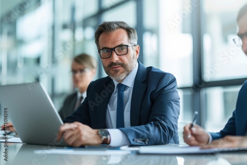 Confident businessman in a suit working on a laptop during a corporate meeting. Professional atmosphere in a modern office setting.