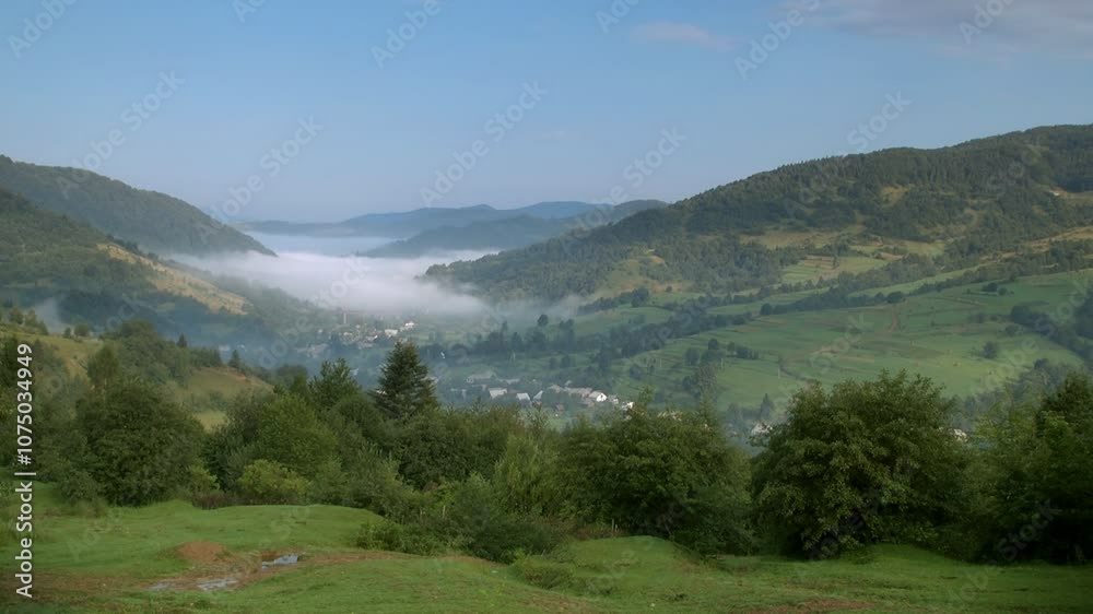 misty valley nestled between green mountains, with a peaceful village and lush forests below. The early morning fog adds a mystical atmosphere, highlighting the serene rural landscape and beauty of un