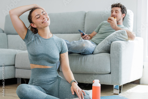 Young sporty woman doing stretching and meditation exercises in her living room while her partner eats a croissant sitting on the sofa, healthy life versus sedentary life, good and bad habits
