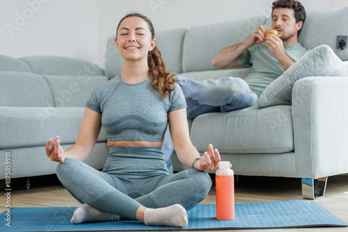 Young sporty woman doing stretching and meditation exercises in her living room while her partner eats a croissant sitting on the sofa, healthy life versus sedentary life, good and bad habits
