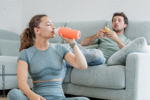 Young sporty woman doing stretching and meditation exercises in her living room while her partner eats a croissant sitting on the sofa, healthy life versus sedentary life, good and bad habits
