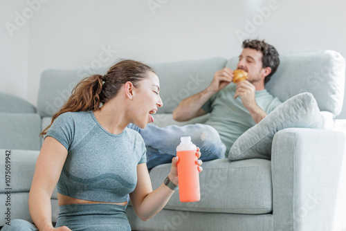 Young athlete exercising in his living room while his partner eats a croissant sitting on the couch, feels shame, anger and disgust healthy life versus sedentary life, good and bad habits
