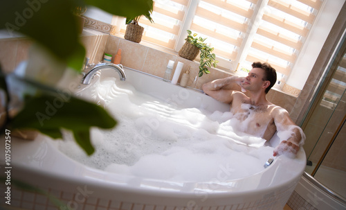 A man enjoys a bubble bath in a bright, modern bathroom. Surrounded by plants and bath products, he looks relaxed, leaning on the edge of the tub as sunlight streams in through the window blinds.