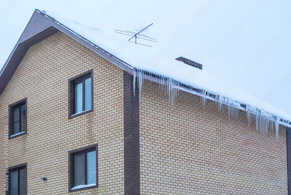 There is snow on the roof of a brick house, and icicles hang from the roof. An antenna is installed on the roof of the country house. The brickwork of the walls of the house. There is snow on the roof of a brick house, and icicles hang from the roof. An antenna is installed on the roof of the country house. The brickwork of the walls of the house.