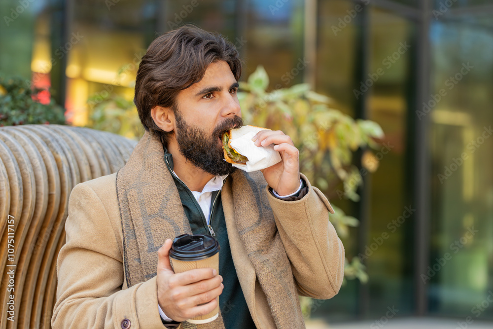 Indian young businessman tourist enjoy lunch break eating tasty sandwich meal and drinking hot beverage coffee or tea. Arabian bearded man guy relaxing sitting on a bench on downtown city street Indian young businessman tourist enjoy lunch break eating tasty sandwich meal and drinking hot beverage coffee or tea. Arabian bearded man guy relaxing sitting on a bench on downtown city street
