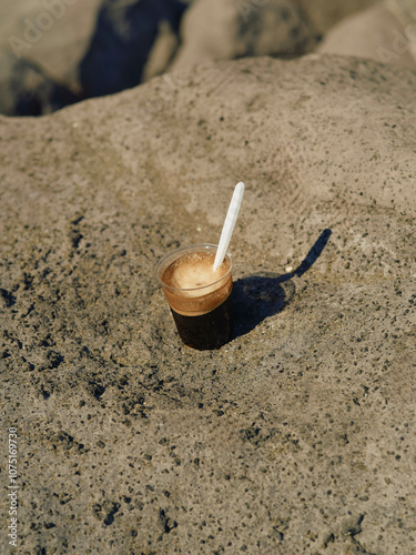 An espresso glass with delicious black coffee and a white plastic spoon standing on rocks near the ocean, Naples, Italy photo