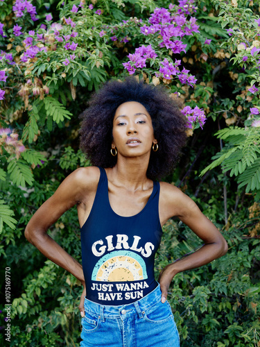 Confident woman with a striking afro stands before a bush of purple flowers, wearing a tank top and blue jeans. photo