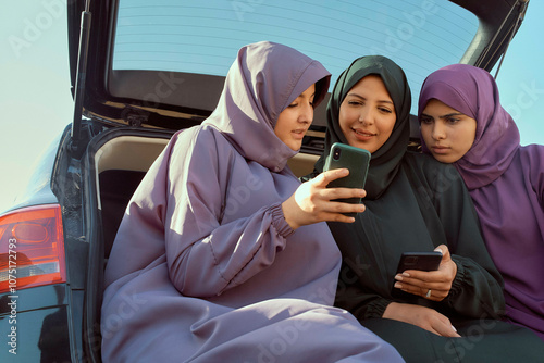 Three women in hijabs looking at a smartphone by a car under a blue sky, Morocco photo