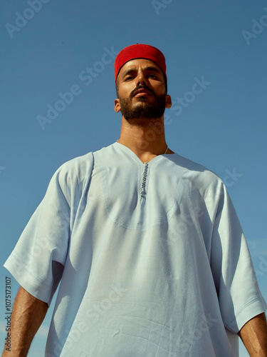 Confident man with a beard wearing a traditional white robe and red headband against a clear blue sky, Morocco photo